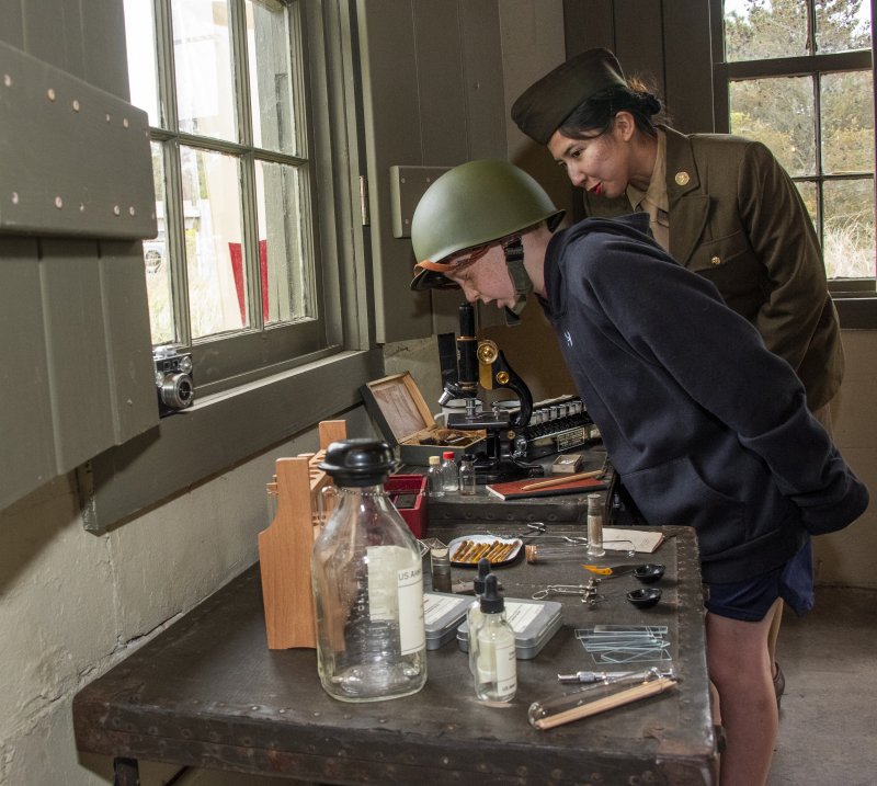 Lo Benitez, a Women’s Army Corps interpreter, explains to James Curtis what to look for to locate a pathogen under a microscope at the Fort Miles Defense Day event.